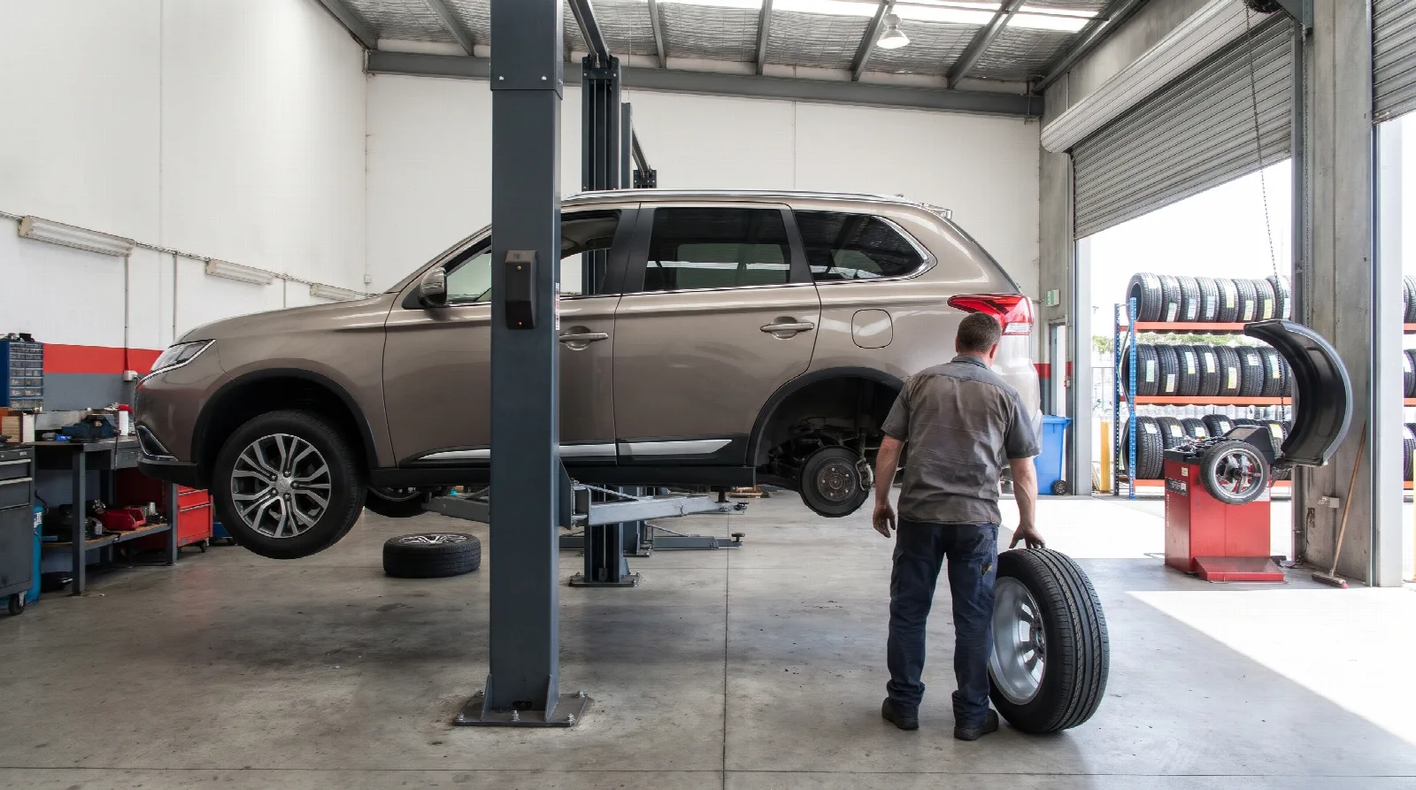 A family SUV on a hoist in a tyre workshop while a mechanic rolls a new wheel towards it